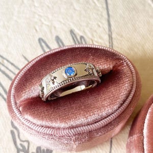Könnte beinhalten: Ein silberner Ring mit einem blauen Edelstein in der Mitte, eingefasst in einem dekorativen Band. Der Ring wird in einer rosa Samt-Ringbox präsentiert. Das Band weist verzierte Details und einen strukturierten Rand auf.