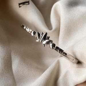 May include: A silver bangle bracelet with a lotus flower design at its centre. The bracelet has a polished finish and decorative patterns along the band. The lotus flower is the focal point, with detailed petals and a smooth, reflective surface.