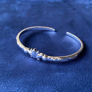 Könnte beinhalten: Ein silbernes Armreif-Armband mit floralem Design. Das Armband hat eine glatte, polierte Oberfläche und ein detailliertes Blumenmuster in der Mitte. Das Armband ist offen und liegt auf einer blauen Oberfläche.