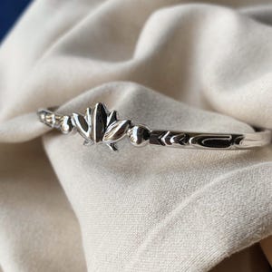 May include: A silver bracelet featuring a lotus flower design. The bracelet has a slender band with decorative details flanking the flower. The bracelet is presented on a white fabric background. The bracelet is approximately 16 cm in circumference.