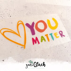 Könnte beinhalten: Ein transparenter Aufkleber mit den Worten "YOU MATTER" in bunten Buchstaben und einer orangefarbenen Herzumrandung. Der Aufkleber hat eine handschriftliche Schriftart und ist wahrscheinlich für Bastelarbeiten oder Dekoration gedacht.