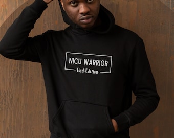 Op de afbeelding: Een zwarte hoodie met een wit rechthoek op de voorkant waarop "NICU WARRIOR - First Edition" staat. De persoon die de hoodie draagt, staat voor een roestige metalen muur.