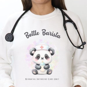 Puede incluir: Sudadera blanca con un oso panda que lleva un gorro de enfermera y sostiene un biberón. El texto "Bottle Barista" está por encima del panda y "Neonatal Intensive Care Unit" está por debajo del panda.