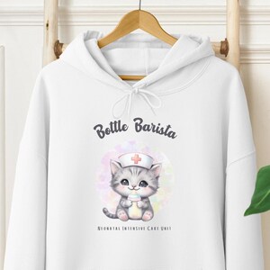 Puede incluir: Sudadera con capucha blanca con un gato gris que lleva un gorro de enfermera y sostiene una botella. El texto "Bottle Barista" y "Neonatal Intensive Care Unit" está impreso en la sudadera.