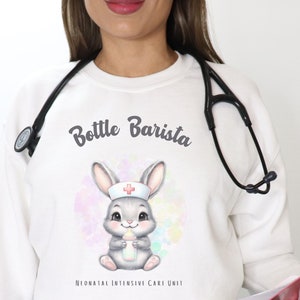 Puede incluir: Sudadera blanca con un conejito gris que lleva un gorro de enfermera y sostiene un biberón. El texto "Bottle Barista" está por encima del conejito y "Neonatal Intensive Care Unit" está por debajo del conejito.