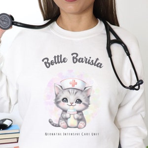 Puede incluir: Sudadera blanca con un lindo gato de dibujos animados que lleva un gorro de enfermera y sostiene una botella. El texto "Bottle Barista" y "Neonatal Intensive Care Unit" está impreso en la sudadera.