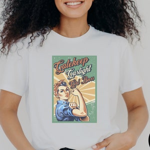 May include: White t-shirt featuring a vintage-style graphic with the words "Gatekeep, Gaslight, Girl Boss" and an illustration of a woman flexing her arm. The design is in shades of blue, red, and yellow.