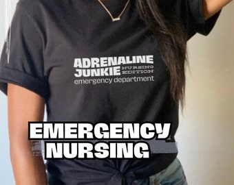 Puede incluir: Una camiseta negra con texto blanco que dice "Adrenaline Junkie Nursing Edition Emergency Department". La persona que lleva la camiseta también lleva un sombrero marrón y unos vaqueros azules.