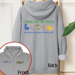 May include: Grey hooded sweatshirt with the text "Caring for Tiny Heroes Pediatric Intensive Care Unit" on the back. The front of the sweatshirt has the text "P.I.C.U." and "Hannah, RN" embroidered in colourful thread.