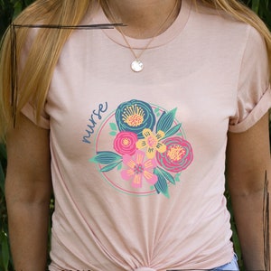 Könnte beinhalten: Ein pinkfarbenes T-Shirt mit einem Blumendesign und dem Wort "nurse" in einem Kreis. Die Blumen sind pink, gelb, blau und grün.