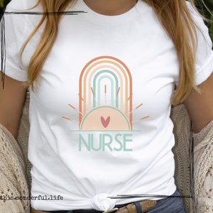 Könnte beinhalten: Weißes T-Shirt mit einem Regenbogen- und Sonnenmotiv und dem Wort "NURSE" in grünem Text.