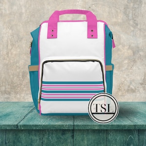 May include: A white backpack with teal and pink stripes. The backpack has a teal zipper and a pink handle. The backpack is sitting on a teal wooden surface. The text "TSL This Wonderful Life" is on a white circle sticker.