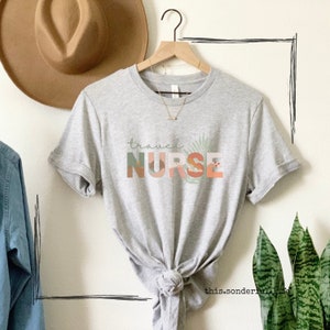 Könnte beinhalten: Graues T-Shirt mit dem Text "travel NURSE" in bunten Blockbuchstaben mit einem Blattdesign.