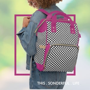 May include: A black and white checkered backpack with pink accents and gold zippers. The backpack has a top handle and adjustable straps. The text "THIS. SONDERFUL. LIFE." is printed below the backpack.