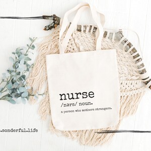 May include: A white tote bag with the word "nurse" and the definition "/nɜːrs/ noun. a person who mothers strangers." printed on it. The bag is lying on a white wooden surface with a piece of greenery and a macrame wall hanging.
