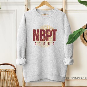 Puede incluir: Sudadera gris con un estampado gráfico en color granate y amarillo que dice "NBPT 01950".