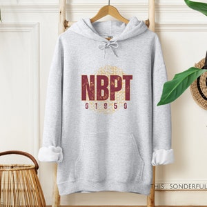Może przedstawiać: Szara bluza z kapturem z nadrukiem graficznym w kolorze bordowym i złotym, na którym widnieje napis "NBPT 01950".