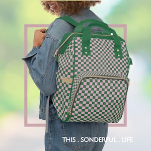 Puede incluir: Una mochila a cuadros verde y rosa con un bolsillo frontal grande. La mochila tiene detalles dorados y cierre de cremallera. El texto "THIS. SONDERFUL. LIFE." está impreso en la parte inferior de la imagen.