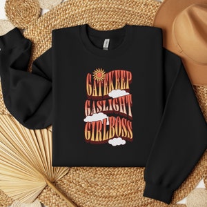 May include: Black sweatshirt with a retro-style graphic design featuring the text "Gatekeep Gaslight Girlboss" in red, orange, and yellow with a sun and clouds.