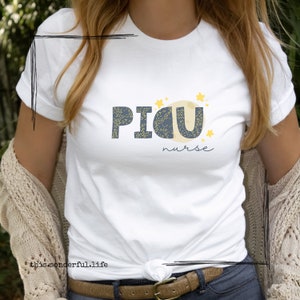 Puede incluir: Camiseta blanca con un gráfico azul y amarillo que dice "PICU nurse" con una luna y estrellas.
