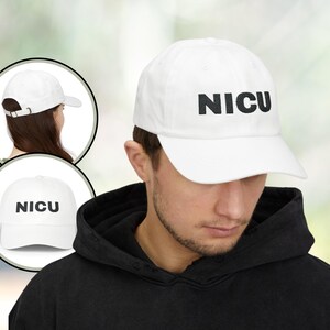 Puede incluir: Gorra de béisbol blanca con la palabra "NICU" impresa en letras negras. La gorra tiene una visera curva y una correa ajustable. La imagen incluye una vista lateral y frontal de la gorra.
