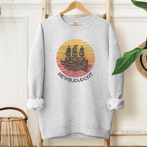 May include: A gray sweatshirt with a vintage-style graphic of a ship sailing on a sunset ocean. The graphic is surrounded by a red, orange, and yellow circle with the text "NEWBURYPORT" below it.