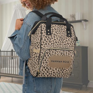 Pode incluir: Mochila para fraldas com estampa de leopardo bege e preta com detalhes em preto. A bolsa tem um bolso frontal e uma etiqueta de nome marrom que diz "HANNAH ROSE". Possui uma alça superior e alças ajustáveis.