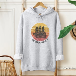 Puede incluir: Sudadera con capucha gris con un gráfico de estilo vintage de un barco navegando hacia una puesta de sol con el texto "NEWBURYPORT" debajo.