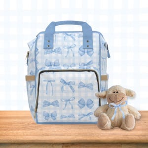 May include: A blue and white checkered backpack with a blue bow pattern. The backpack has a large zippered pocket on the front and two smaller pockets on the sides. A small plush white lamb with a blue bow is sitting in front of the backpack.