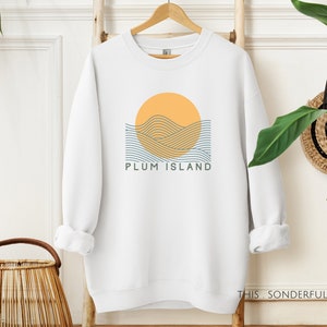 Könnte beinhalten: Weißer Pullover mit einem Grafikdesign einer gelben Sonne, die über blauen Wellen untergeht. Der Text "PLUM ISLAND" ist unter der Grafik gedruckt.