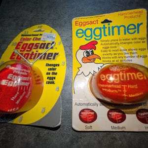 May include: Two Eggsact eggtimer devices in original yellow blister packaging. The left oval card features a clock face design, while the right rectangular card displays a cartoon chicken illustration. Both red timers show soft, medium, and hard settings.