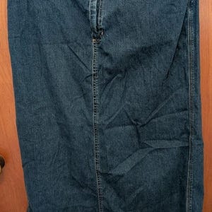 May include: A long, dark-wash denim skirt with a classic five-pocket design. The skirt features a button and zip closure, belt loops, and a straight silhouette. The skirt is hanging on a hanger against a wooden door.