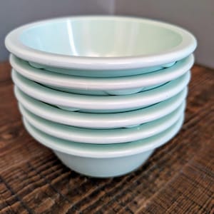 May include: A stack of six light blue ceramic bowls. The bowls are stacked on top of each other and are sitting on a wooden surface.