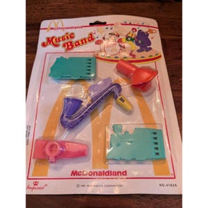 May include: A sealed package of McDonaldland Music Band toys. The toys include a purple saxophone, a red megaphone, a pink whistle, and two green musical instruments. The packaging has the McDonald's logo and the words "Music Band".