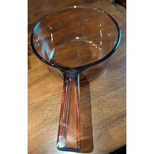 Peut inclure: Une casserole en verre transparent avec une seule poignée. La poignée et le bord ont une légère teinte ambrée. La casserole est vide et repose sur une surface en bois. La casserole est conçue pour la cuisine.
