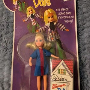 Puede incluir: Un juguete vintage Miss Matchbox Doll en un blíster de plástico. La muñeca es una niña rubia que lleva un vestido azul y zapatos rojos. La muñeca sostiene una pequeña casa. El embalaje es morado y presenta el texto "Miss Matchbox Dolls" y "¡Ella duerme acurrucada y sale a jugar!" La muñeca está etiquetada como "15 - Sally Stewardess".