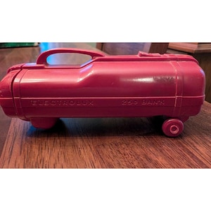 Può includere: Un aspirapolvere giocattolo vintage in plastica rossa con la scritta "ELECTROLUX 25¢ BANK". Il giocattolo ha una maniglia, ruote e un corpo cilindrico. È un oggetto da collezione retrò.