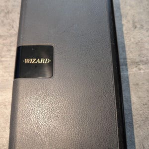 May include: A rectangular electronic organizer with a dark gray textured exterior. The device features a smooth black panel on the left side with the word WIZARD printed in gold lettering. The overall design is compact with rounded corners.