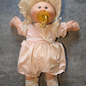 Pode incluir: Uma boneca com rosto redondo, olhos castanhos e cabelo de lã loira, vestindo um vestido rosa pálido com um pequeno padrão floral, uma touca de renda branca e meias brancas. A boneca tem uma chupeta de plástico amarela na boca.