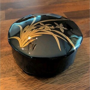 May include: A round, black lacquered box with a glossy finish. The lid features a gold-colored illustration of long, slender leaves and delicate orchid-like flowers extending across the surface. The container has a smooth, reflective appearance.