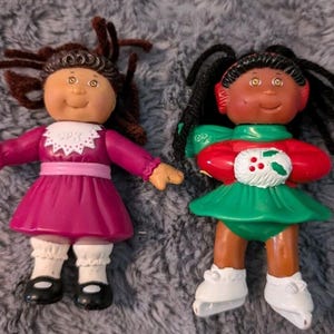 Puede incluir: Dos muñecas Cabbage Patch Kids con atuendos de invierno. Una muñeca tiene cabello castaño y un vestido rosa, mientras que la otra muñeca tiene cabello negro y un vestido verde con una chaqueta roja. Ambas muñecas llevan patines de hielo blancos.