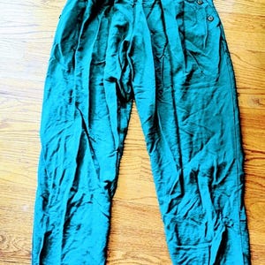 May include: Teal-colored high-waisted pants with pleats and button details. The pants have a relaxed fit and are made of a slightly shiny fabric. The buttons are visible on the waistband and along the side of the pants.