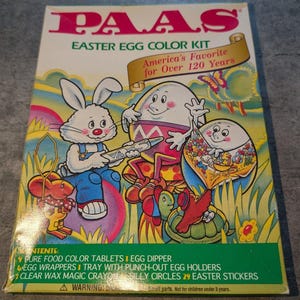 Puede incluir: Una caja de kit para colorear huevos de Pascua PAAS con un conejo blanco, una tortuga y dos personajes de huevo en una escena colorida. El texto dice PAAS EASTER EGG COLOR KIT, America's Favorite for Over 120 Years, y lista el contenido.