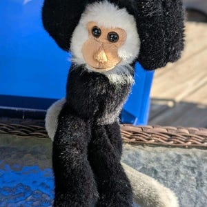 May include: A black and white plush monkey toy with a long tail hanging from a blue surface. The monkey has a white face with black eyes and a brown nose.