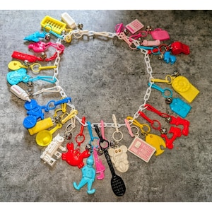 Puede incluir: Un collar de cadena de plástico blanco con un surtido de dijes en miniatura coloridos, que incluyen una bicicleta azul, un cangrejo rojo, un teléfono amarillo, un biberón blanco, una raqueta de tenis negra y otras formas pequeñas con cascabeles.