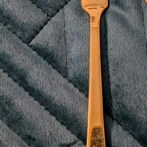 Könnte beinhalten: Ein goldenes Buttermesser mit abgerundeter Klinge und einem dekorativen Griff. Das Messer liegt auf einem blauen Stoffhintergrund.