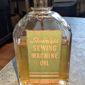 May include: A clear glass bottle filled with amber-coloured liquid, featuring a black ribbed screw cap. A rectangular green label on the front displays the text "Stainless SEWING MACHINE OIL" in a light-coloured, sans-serif and script font.