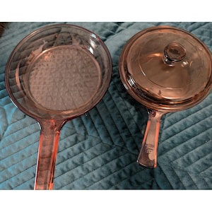May include: Two pieces of pink-tinted glass cookware. One is an open skillet with a textured diamond pattern on the base. The other is a saucepan with a matching glass lid featuring a central knob handle. Both have long, flat glass handles.