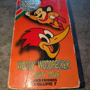 May include: A rectangular VHS tape case with a bright orange background featuring illustrations of Mighty Mouse and Woody Woodpecker. The text reads Woody Woodpecker & Mighty Mouse and Friends Volume 7. A silver circular sticker is at the top left.