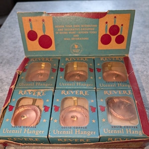 May include: A vintage cardboard display box containing six individual Revere Ware solid copper utensil hangers. Each box features a blue and red design with the text 'REVERE SOLID COPPER Utensil Hanger' and illustrations of hanging kitchen tools.